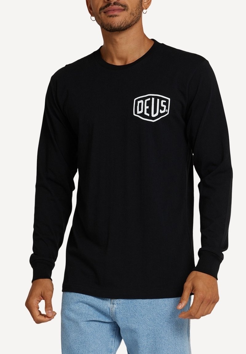 Schwarzes Langarm-T-Shirt aus Baumwolle mit einem weißen DEUS-Logo in Schildform auf der linken Brustseite. Klassisches Fit-Design.