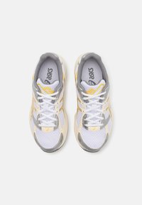 Une paire de chaussures de course Asics blanches et jaunes avec des accents gris et des lacets blancs, vue de dessus sur un fond clair.