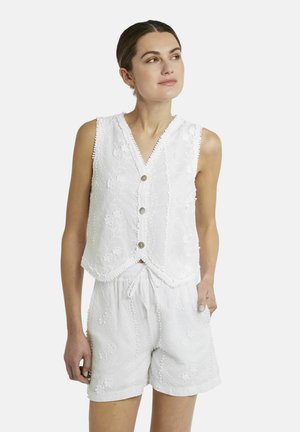 Frau trägt weiße ärmellose Weste mit Knöpfen und passende bestickte Shorts, steht mit einer Hand in der Tasche und schaut zur Seite.