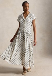 Nainen pukeutuneena kermaankukkaiseen, lyhythihaiseen mekkoon, jossa on röyhelöt, seisoo ja pitää mekonsa helmaa kevyesti kiinni katsellen sivulle.