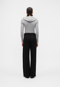 Personne aux cheveux tressés portant un sweat à capuche gris et un pantalon noir à jambes larges, debout de dos sur un fond uni.
