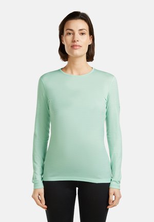 Kobieta o krótkich ciemnych włosach, ubrana w jasnoliemniową bluzkę z długim rękawem i czarne spodnie, stojąca na tle jednolitego białego tła.