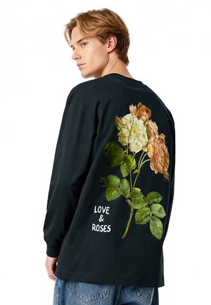 Junger Mann in schwarzem Langarmshirt mit großem Blumenmuster und dem Schriftzug „LOVE & ROSES“ auf dem Rücken, kombiniert mit blauen Jeans.