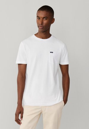 Mann trägt ein schlichtes weißes T-Shirt mit kurzen Ärmeln und Brusttasche sowie hellbeige Hose vor einfarbigem grauen Hintergrund.