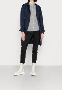 Trench-coat bleu marine à double boutonnage, porté sur une chemise rayée, associé à un pantalon noir et des bottines blanches à semelle épaisse.