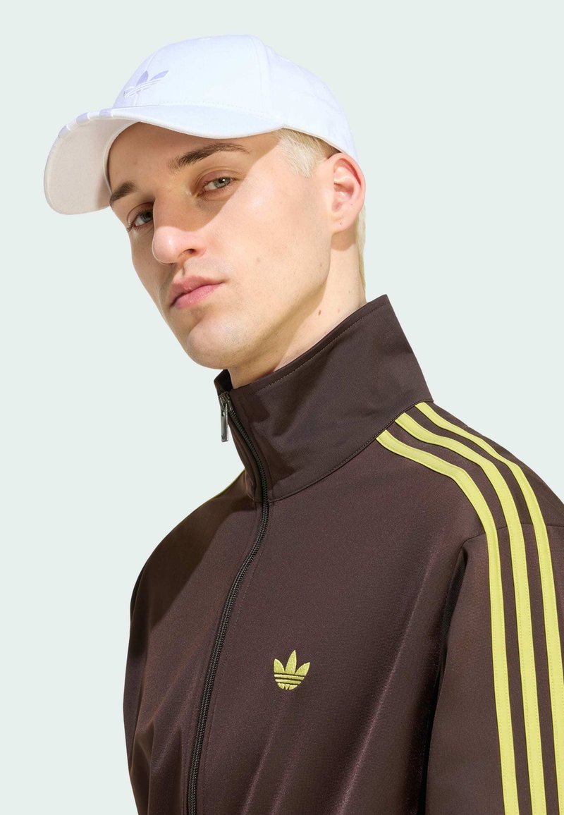 Giovane uomo dalla pelle chiara e capelli biondi corti, indossa un berretto bianco e una giacca marrone con zip con strisce gialle sulle spalle.