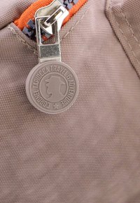 Tela beige con una superficie texturizada que presenta una cremallera naranja. Una etiqueta redonda de color gris claro muestra una silueta y texto. Incluye herrajes plateados.
