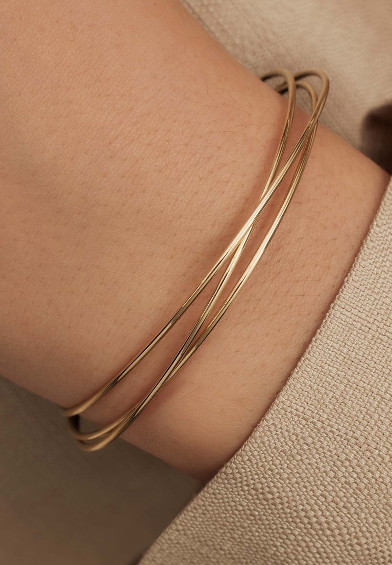 Triple gold-tone metal bangle bracelet, featuring a sleek, smooth design with intertwining strands and a polished finish on a wrist.