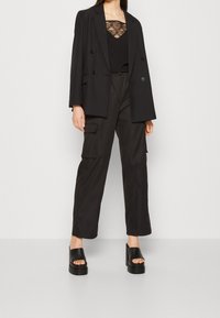 Czarna marynarka z podwójnymi guzikami, w połączeniu z czarną koronkową kamizelką, luźnymi czarnymi spodniami cargo oraz masywnymi czarnymi sandałami na platformie.