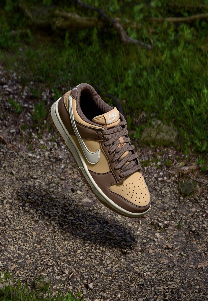 Baskets Nike marron et beige avec un swoosh blanc flottant au-dessus d’un chemin de gravier avec de l’herbe verte en arrière-plan.