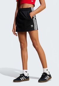 Črna športna krilo s tremi belimi stranskimi črtami, elastičnim pasom in žepi. Kombinirana z rdečo kratko majico in črnimi supergami.