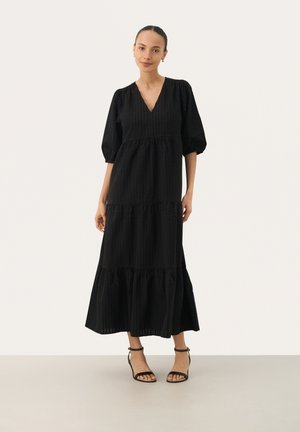 Svart långklänning med lagerdesign, V-ringning och puffärmar. Tillverkad av textil med struktur, tillsammans med svarta sandaletter med remmar.