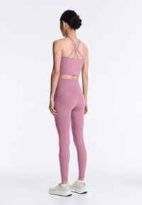Femme debout, de dos, portant un crop top mauve à bretelles croisées et des leggings taille haute assortis avec des baskets beiges sur fond blanc.