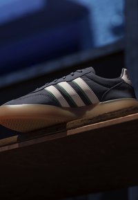 Zapatilla deportiva azul oscuro con rayas blancas y verdes y suela marrón claro descansando sobre una superficie de madera con fondo desenfocado.