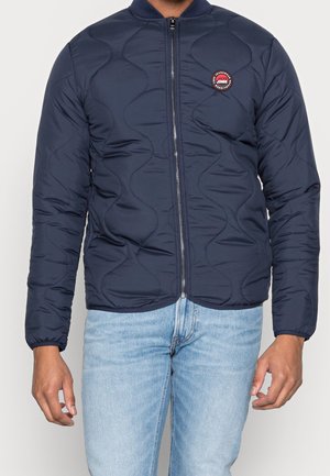 Homme portant une veste matelassée bleu marine avec fermeture éclair et logo Jack & Jones sur la poitrine, associée à un jean bleu clair.