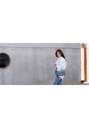 Zelená kožená crossbody taška s plochým silhouette a popruhem. Taška má minimalistický design a decentní logo akcent. Pozadí tvoří beton.
