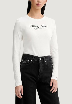 Biała koszula z długim rękawem z napisem "Tommy Jeans" w czarnym kursywie. Do tego czarne jeansy z wysokim stanem, z przodu z guzikami i łatą.