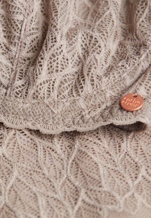 Lace-patterned fabric in light beige, featuring a scalloped edge and a small copper-tone logo tag, highlighting intricate textural detail.