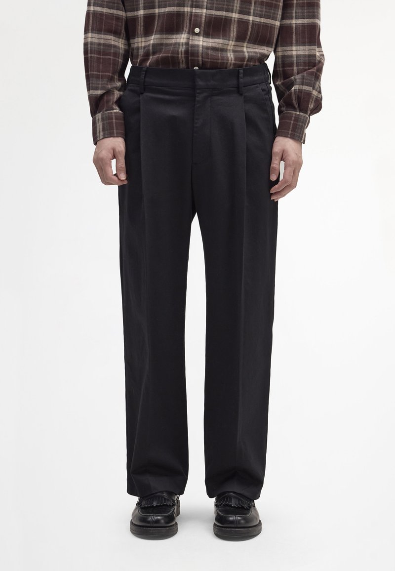 Schwarze plissierte Hose aus Baumwolle mit lockerer Passform und Seitentaschen, kombiniert mit schwarzen Lederschuhen und einem braunen karierten Hemd.