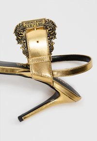 Gold metallic high-heeled sandal with a textured finish and intricate lion head buckle. Features a black insole and slim strap design.