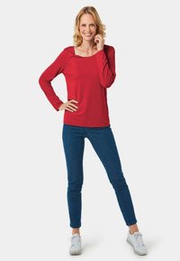 Rotes Langarmoberteil mit quadratischem Ausschnitt, kombiniert mit blauen Slim-Fit-Jeans und weißen Sneakers. Glatte Textur, minimalistisches Design.