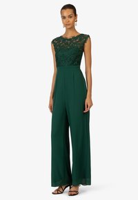 Green jumpsuit with lace bodice and cap sleeves, wide-leg pants, pleats, and a smooth fabric finish. Black strappy sandals complete the look.