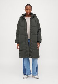 Grønn polstret jakke med hette, knapp- og glidelås-lukking, sidelommer og kne-lang design. Brukt over en lett genser og jeans.