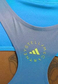 Svijetloplava sportska grudnjak s dizajnom na leđima. Ima zeleni kružni logotip s tekstom i tri pruge u sredini. Glatka tekstura.