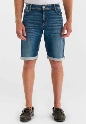 Mid-lange, opgerolde denim shorts voor heren, gedragen met bruine leren veterschoenen op een lichte achtergrond.