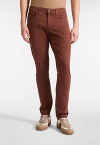 Brązowe bawełniane spodnie o slim fit, pięć kieszeni, z metalowym zapięciem i subtelną teksturą. Połączone z beżowymi i brązowymi trampkami.