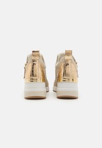 Sneakers beige con accenti metallici dorati e superficie testurizzata. Presentano un anello posteriore e una suola bianca spessa con fondo beige.