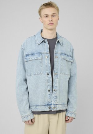 Jeune homme aux cheveux blonds courts portant une veste en denim bleu clair sur une chemise sombre et un pantalon beige, debout devant un fond gris.