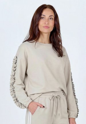 Kvinde med langt brunt hår iført en beige sweatshirt med flæseærmer og matchende bukser med snøre, stående med den ene hånd i lommen.