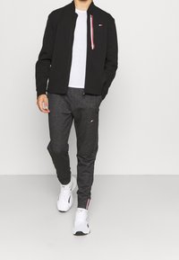 Schwarze Zip-Jacke mit Stehkragen und Akzenten in Rot, Weiß und Blau. Graue, schmal zulaufende Jogginghosen mit kleinem Logo. Weiße Sneakers.