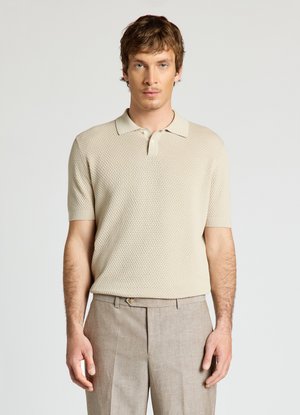 Uomo che indossa una polo beige a maniche corte con trama e pantaloni marrone chiaro, in piedi davanti a uno sfondo chiaro e uniforme.
