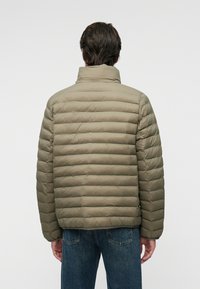 Paminkštintas alyvuogių žalia striukė su aukštu apykakle, su horizontaliu siuvinėjimo dizainu ir elastinėmis rankovėmis, dėvima virš tamsių džinsų.