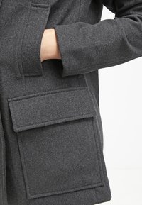 Dunkelgrauer Wollmantel mit großer Klappentasche und sichtbarer Hand darin. Der Stoff hat eine strukturierte Oberfläche und ein gerades, strukturiertes Design.