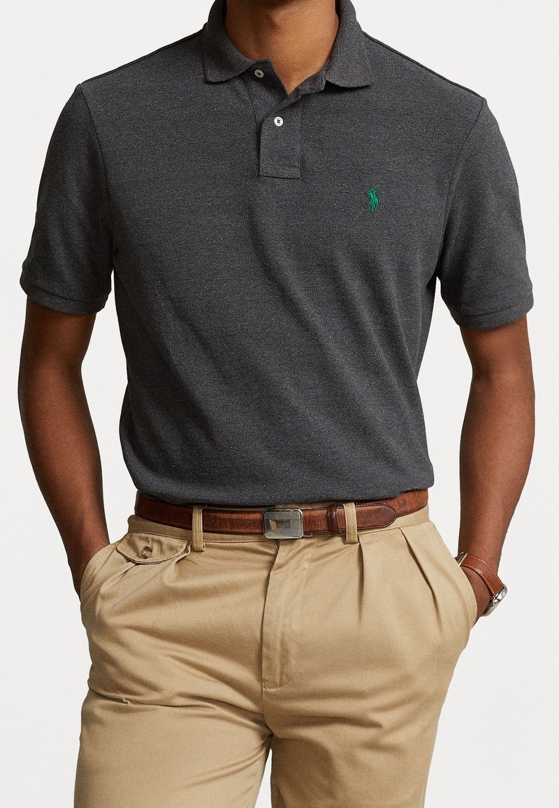 Homme portant un polo à manches courtes gris foncé avec un logo vert, un pantalon plissé beige, une ceinture en cuir marron et une montre-bracelet, les mains dans les poches.