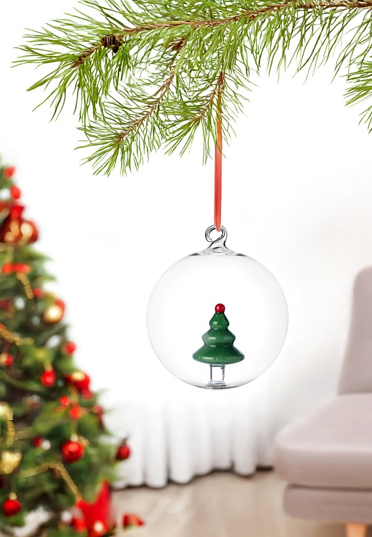 Szklana ozdoba choinkowa z figurką zielonej choinki w środku, zawieszona na czerwonej wstążce, wisząca z gałęzi sosny.
