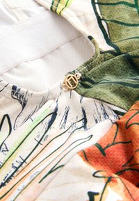 Gros plan d’un tissu floral multicolore avec une boucle circulaire dorée et une doublure en mesh blanche au niveau de l’encolure du vêtement.