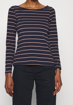 Kvinde iført navyblå langærmet skjorte med tynde vandrette beige striber og sorte bukser, står foran en ensfarvet baggrund.