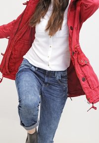 Rode parka met trekkoorden en knopen, witte overhemden en opgerolde blauwe jeans. Zwarte enkelaars maken de look compleet.