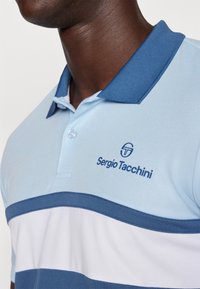 Ljusblå polo med mörkblå krage, horisontella ränder i marinblått och vitt, samt en broderad "Sergio Tacchini" logotyp på bröstet.