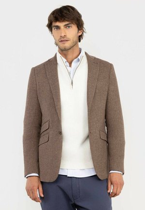 Hombre con blazer marrón texturizado sobre un suéter blanco con cremallera y camisa azul claro con cuello, de pie frente a un fondo sencillo.