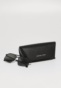 Occhiali da sole neri con montatura geometrica, lenti scure e accenti argento, abbinati a un astuccio nero testurizzato con il logo "MICHAEL KORS".