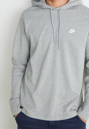Person iført en lysegrå Nike hættetrøje med snore og marineblå bukser, hænder i lommerne, mod en ensfarvet hvid baggrund.