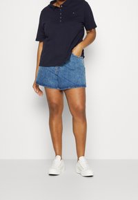Marinblå pikétröja med korta ärmar och knappslå, ihop med denimshorts och vita sneakers, ståendes mot en enfärgad bakgrund.