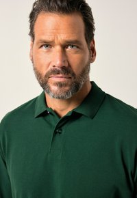 Polo verde de algodón, con cuello, tres botones y una textura suave. El modelo tiene barba y cabello corto y oscuro.