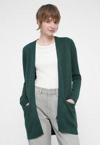 Cardigan verde scuro con apertura frontale, texture a maglia, polsini a costine e due tasche davanti. Abbinato a una maglietta bianca e pantaloni grigio chiaro.