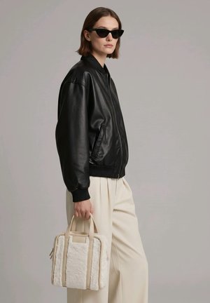 Mujer con chaqueta de cuero negra, gafas de sol negras estilo cat-eye, pantalones anchos color crema, sosteniendo un bolso beige texturizado sobre fondo gris.
